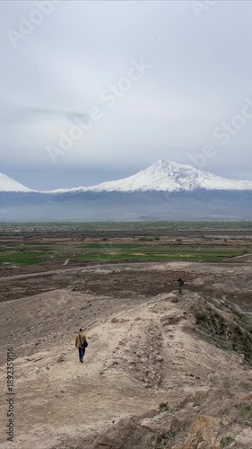 Hiker walking on a rocky trail with majestic snow-capped Mount Ararat in the distance under an overcast sky, overlooking a vast green valley.
