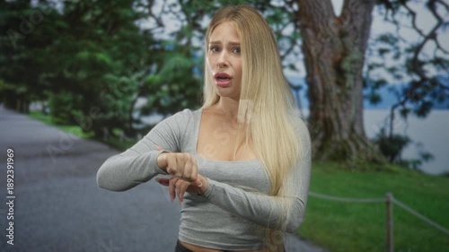 Woman checks wrist and points to her wrist in park, gray longsleeve crop top, hands raised in questioning shrug; waiting lateness anxiety.