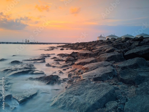 A beautiful, warm-toned photograph of a rocky shore as the sun sets or rises over the ocean. The sky is filled with soft orange, pink, and blue hues, reflecting gently on the water