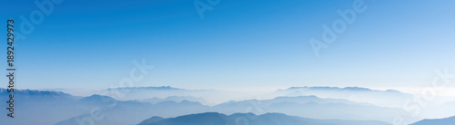 Panoramic view of majestic mountain ranges layered in mist under a clear blue sky. A peaceful and serene natural landscape capturing the vast horizon of the mountains.