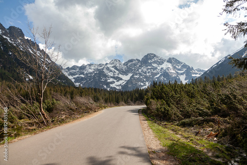 Scenic view of Tatra Mountains in Poland with a winding road, lush greenery, and snow-capped peaks under a partly cloudy sky