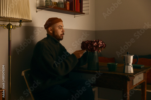 Φωτογραφία A contemplative man in a green turtleneck and orange beanie sitting at a wooden table, looking at purple flowers in a cozy, dimly lit interior with a vintage atmosphere