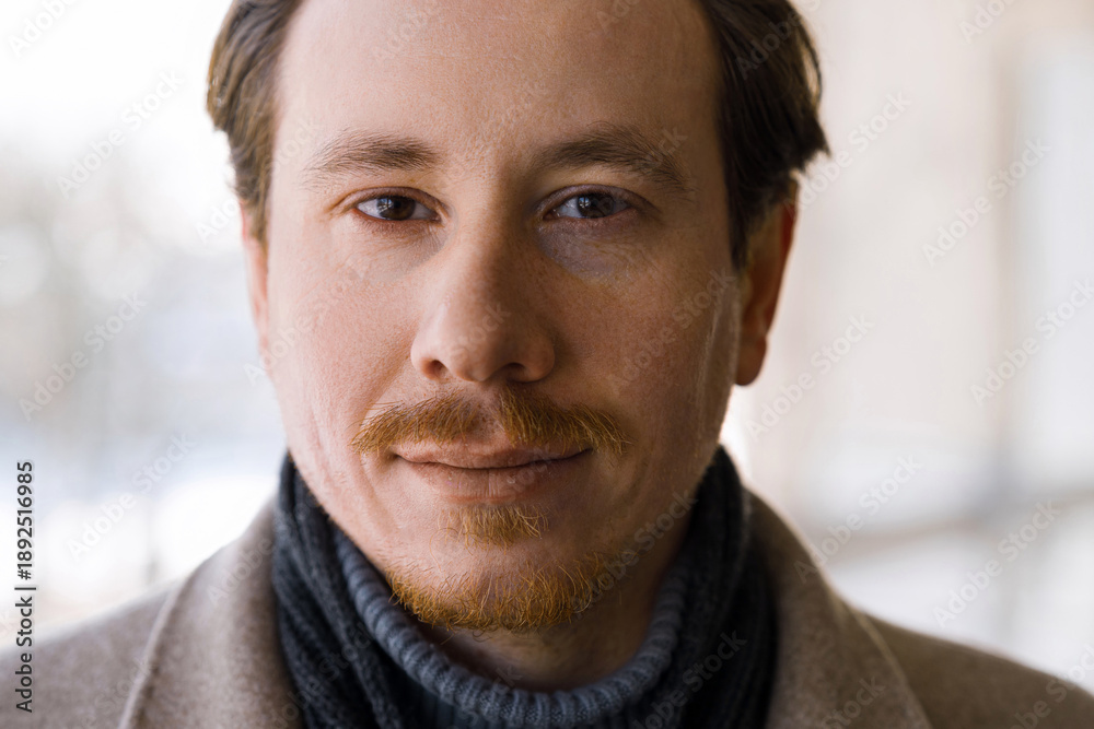 Fototapeta premium Close-up portrait of an attractive young redhead caucasian man wearing a coat on a cold, snowy winter day while walking down the street.