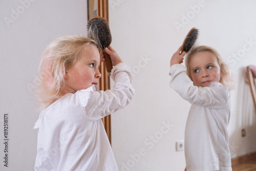 Cute little girl brushing her hair in front of the mirror at home.