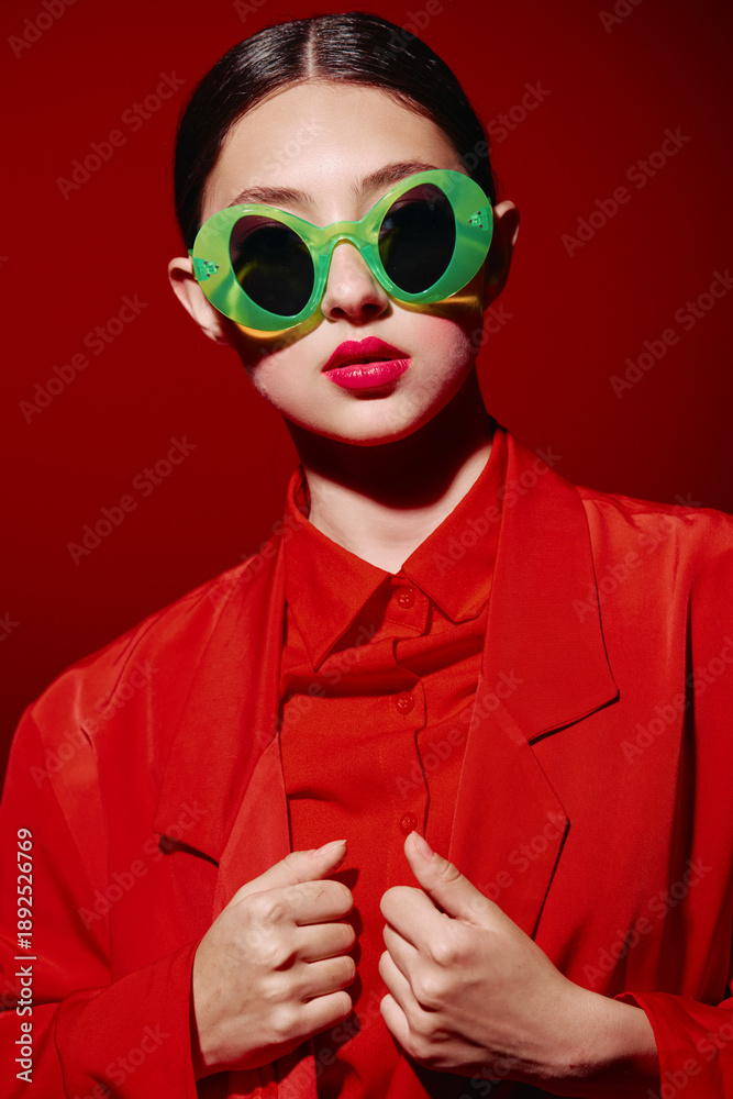 Fototapeta premium Fashionable woman in vibrant green round sunglasses and bold red lips poses in a red outfit on red background. Creative beauty and high fashion concept with striking colors.