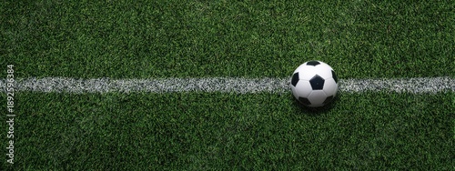 A close-up view of a soccer ball on a green grass field with a white line running across the center. The ball rests on the right