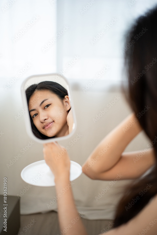Naklejka premium Beautiful asian woman touching her smooth glowing skin with closed eyes, smiling in mirror reflection. Confident girl proud of natural beauty and healthy skincare results.