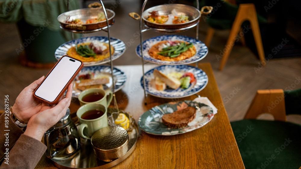 Fototapeta premium Person holding a smartphone while enjoying a tiered tray of food and tea