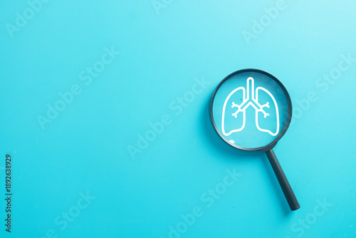 Magnifying glass focusing on white human lungs icon on blue background with copy space. Respiratory health checkup, lung cancer diagnosis, pneumonia virus, and air pollution protection concept.