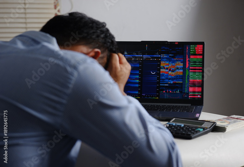 Rear view of a shocked and stressed businessman, a frustrated young man wearing formal clothes, lowering his head while sitting in front of a computer with business charts and falling stocks
