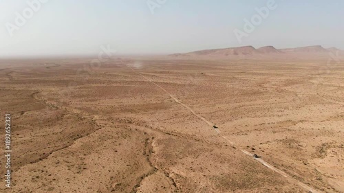 Wide drone shot showing offroad vehicles traveling along long tracks across an expansive arid terrain highlighting isolation scale and the immensity of the landscape