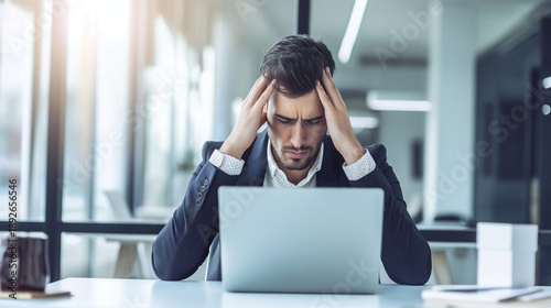 Stressed businessman holding his head in frustration while looking at his laptop in an office setting, symbolizing work pressure and deadlines