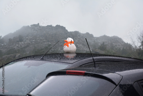 Little snowman on top of a parked car roof, with snowy mountains in the background, winter and cold