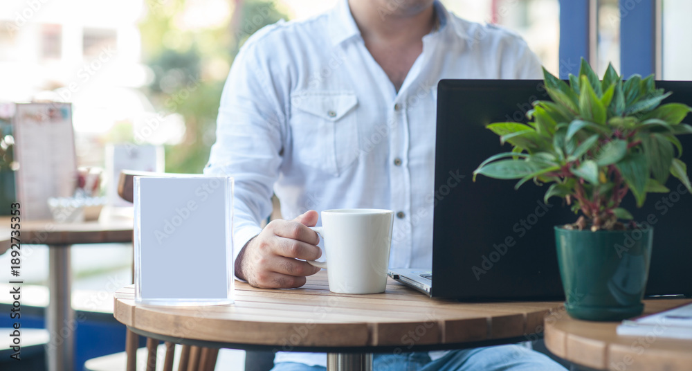 Fototapeta premium A white coffee mug beside a laptop on a wooden café table, with a person softly out of focus in the background, creating a calm modern workspace and lifestyle atmosphere.
