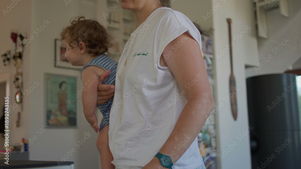 Fototapeta premium Mother gets up from chair and lifts baby in arms, mid-motion action in bright home interior filled with books, photos, and everyday domestic warmth