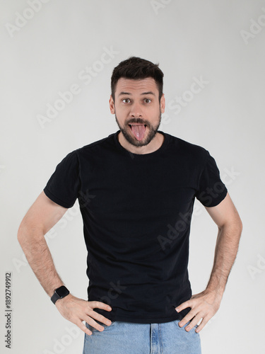 Studio portrait of a charismatic millennial man with a beard, in a basic black t-shirt and blue jeans. He is making a funny, playful facial expression, sticking his tongue out.