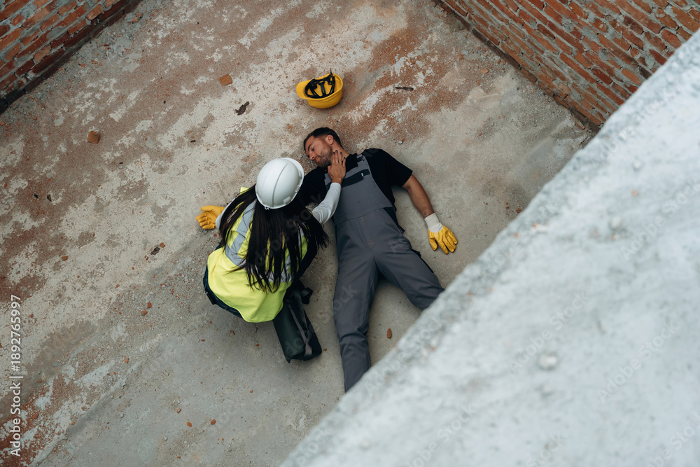Fototapeta premium Top view. Man, construction worker fell from big height, lying down without consciousness, accident, woman is checking
