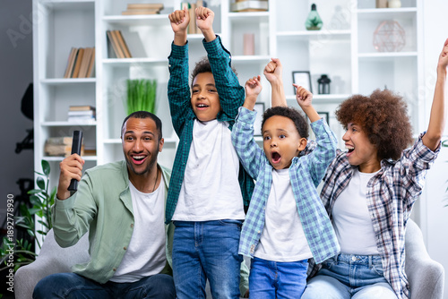 Low angle front view of four members family at home sitting on a sofa in the living room, watching TV and all cheering and rasing fists in victory