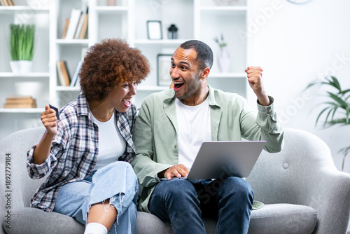 Joyful sincere laughing young african american family couple celebrating internet success, getting online lottery giveaway gambling auction win notification, feeling excited looking at laptop screen.