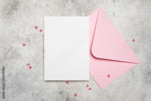 Blank greeting or invitation card mockup with pink envelope and small hearts decoration on grey stony background