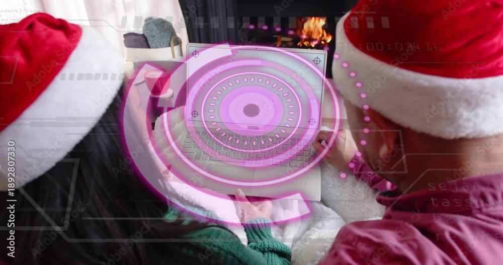 Fototapeta premium Holding couple wearing Santa hats sharing laptop on couch, displaying magenta holo UI and fireplace