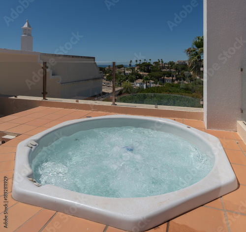 A hot tub sits on a rooftop terrace, surrounded by clear sky and palm trees, offering a view of the ocean and nearby landscape in bright weather