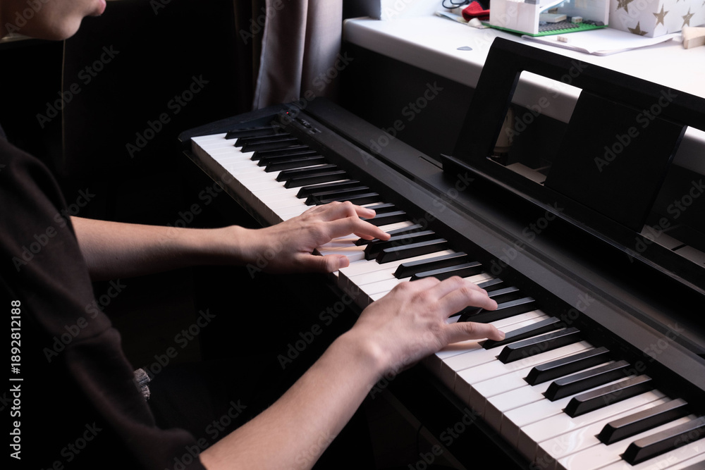 Fototapeta premium A pair of hands plays a sleek black piano keyboard. Soft lighting highlights the fingers pressing the white and black keys, creating a moody, artistic atmosphere. Close-up shot, dark background.