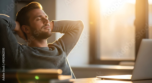 Relaxed young man sitting at his desk with laptop leaning back with eyes closed in warm sunset light. Lifestyle photography.