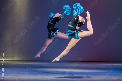 Tableau sur toile Two young cheerleaders perform a synchronized jump with high leg extensions, holding blue pom-poms mid-air
