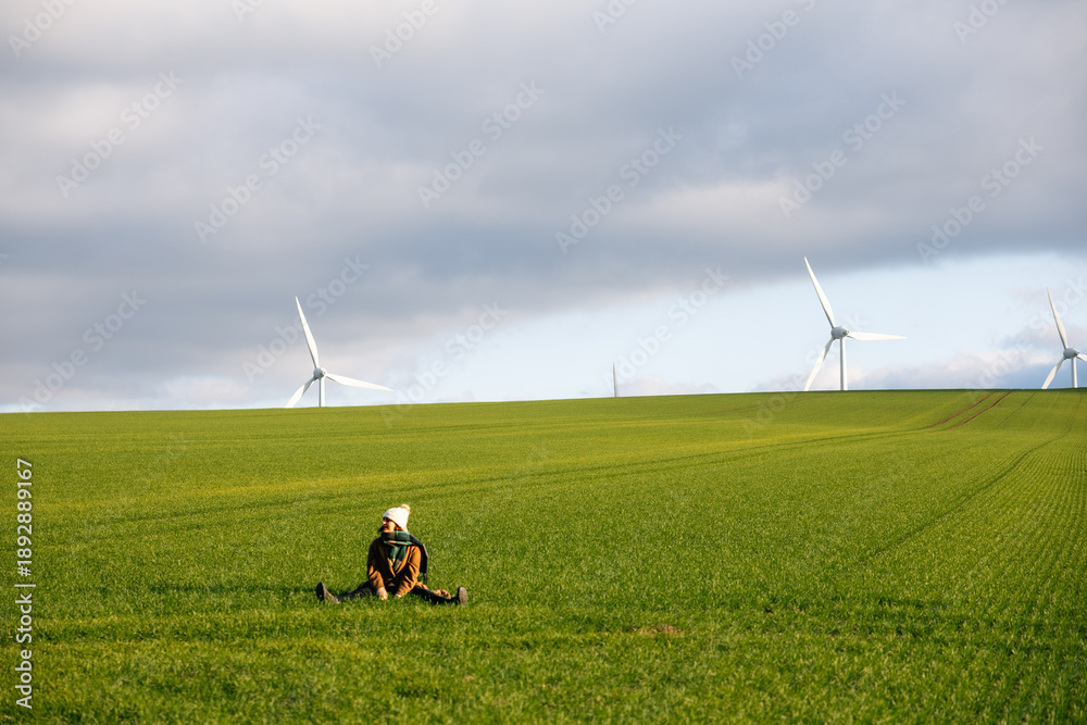 Fototapeta premium Young Woman Relaxing in Green Meadow with Wind Turbines in Background