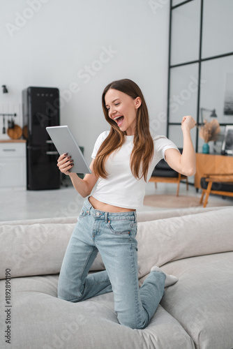 Excited woman looking at tablet screen, celebrating success, showing yes gesture, sitting on couch, smiling overjoyed young female received good news in message, online lottery win. Vertical shot