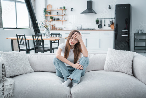 Young tired unhealthy Caucasian woman sitting on couch in living room at home holding head with hand suffering from strong sudden headache or migraine throbbing pain.
