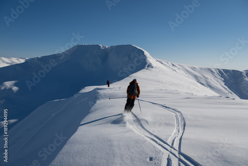 Off-piste freeride skiers traverse a snow-covered ridge under a clear blue sky in fresh powder on a high alpine peak
