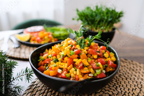Fresh colorful corn salad with cherry tomatoes, bell pepper, green onions and herbs served in a black bowl on a rustic table. Healthy vegan summer side dish with vibrant vegetables and natural light.