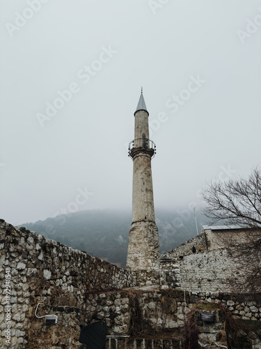 Foto Tall stone minaret standing inside the walls of Travnik Fortress, surrounded by weathered stone and misty mountain landscape