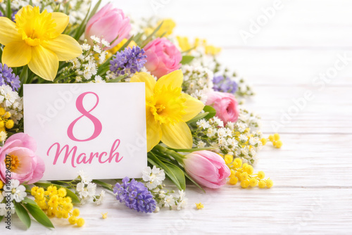 Greeting card with March 8 text on a bouquet of spring flowers like daffodils, tulips, and mimosa on white wood.