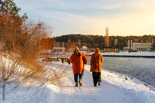 Two people in warm winter coats walking along a snowy path by a river at sunset. Golden hour lighting on a peaceful winter landscape with bare bushes and a forest in the background.
