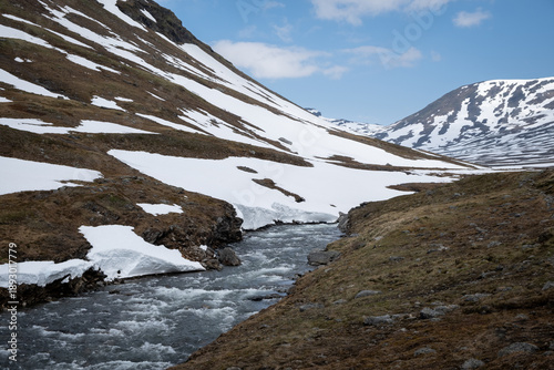 Cold mountain river rushes through a narrow valley between snow covered slopes along the Kungsleden trail in Swedish Lapland