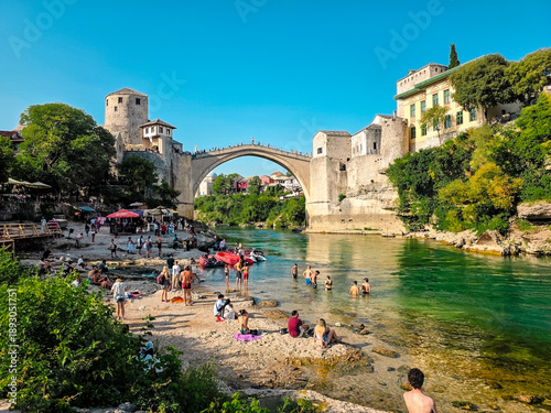 Vászonkép Old town of Mostar with historic stone houses, traditional bridge and turquoise river surrounded by green hills and mountains under clear summer sky