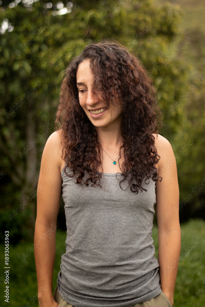 Naklejka premium Portrait of a cheerful female with natural look standing outdoors with a green blurred background, looking down with a shy and genuine smile.