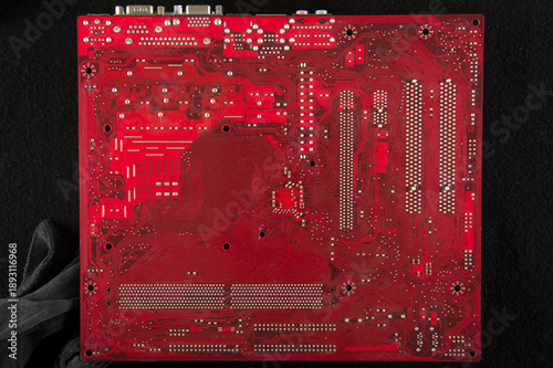 Red printed circuit board back side with solder points and traces on dark background, electronics motherboard texture