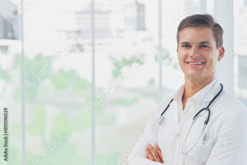 Foto Male doctor posing arms-crossed smiling at clinic windows in lab coat, stethosco
