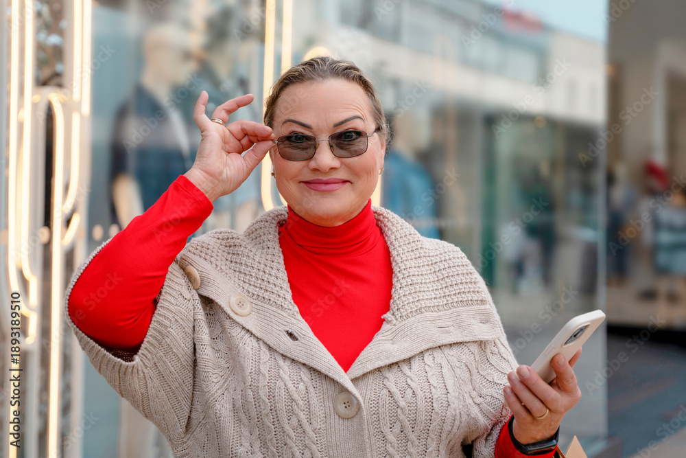 Fototapeta premium Woman wearing sunglasses holds a smartphone while smiling outside a shopping area in city