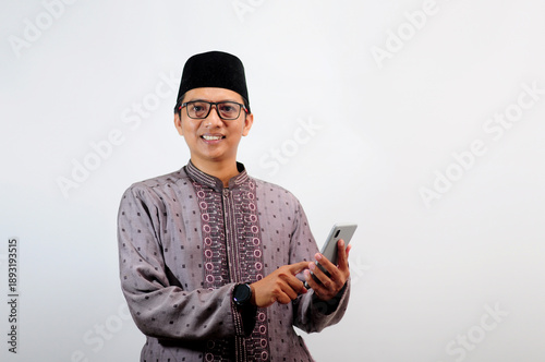 An Asian Muslim man smiles happily while sending a message using his mobile phone, isolated on white background