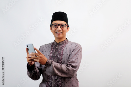 An Asian Muslim man smiles happily while sending a message using his mobile phone, isolated on white background
