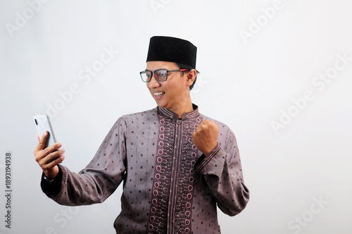 An Asian Muslim man smiles happily while sending a message using his mobile phone, isolated on white background