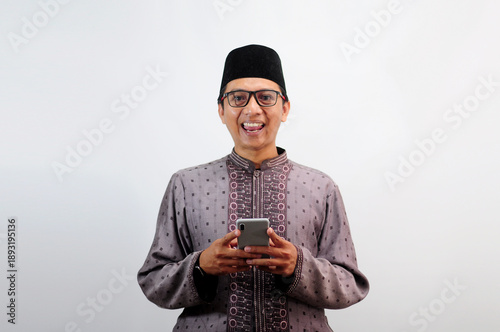 An Asian Muslim man smiles happily while sending a message using his mobile phone, isolated on white background