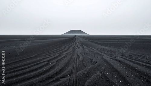 A vast black sand beach with tire tracks stretching towards a distant island hill under a gray sky
