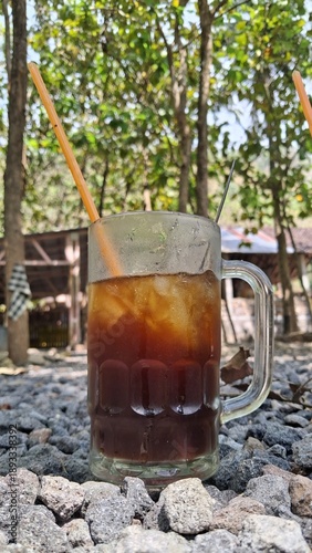 Iced coffee in a clear glass mug served outdoors with natural daylight. Refreshing drink concept, rustic setting, casual lifestyle, summer refreshment, minimal composition with natural background