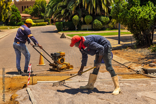 Two african american construction workers leveling gravel during road paving, vibratory plate compactor